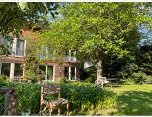 Holzhaus mit großen Fenstern und Balkonen, umgeben von einem Garten mit grünen Pflanzen, einem Baum und Sitzgelegenheiten. Im Vordergrund stehen ein hölzerner Stuhl und ein Tisch, umgeben von Gras und Blumen.