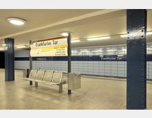 U-Bahn-Station "Frankfurter Tor" in Berlin mit blau gefliesten Wänden, Metallsäulen und einer Sitzbank aus Metall. Ein Hinweisschild mit Linien- und Ausgangsinformationen ist zentral platziert.