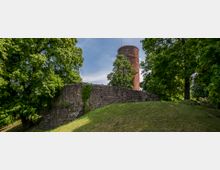 Ruine einer historischen Steinmauer mit einem runden Backsteinturm im Hintergrund, umgeben von dichtem Grün und hohen Bäumen, auf einem leicht hügeligen Grasgelände.