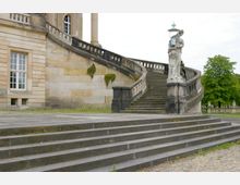 Historisches Gebäude mit einer geschwungenen Steintreppe, verziert mit Balustraden und einer Figurengruppe, die eine Laterne trägt. Im Hintergrund sind Bäume und ein steinerner Zaun sichtbar.