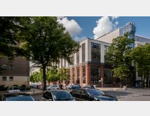 Straßenszene mit einem großen, modernen Bürogebäude aus Glas, Stahl und rotem Backstein, umgeben von Bäumen. Im Vordergrund sind parkende Autos, eine Kreuzung und Fußgänger zu sehen.