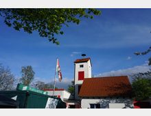 Weißes Gebäude mit rotem Ziegeldach und einem kleinen Turm mit roten Fensterläden, daneben eine Fahne auf einem Mast und umgeben von Bäumen unter einem klaren blauen Himmel. Ein grüner Container mit der Aufschrift „Wernesgrüner“ steht im Vordergrund.