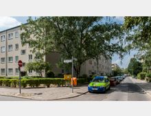 Straßenansicht einer Wohngegend mit einem mehrstöckigen beige-grauen Wohngebäude links, umgeben von Bäumen und gepflegten Hecken. Am Straßenrand stehen parkende Autos, ein Straßenschild mit der Aufschrift "Wilseder Straße", ein Briefkasten und ein Abfallbehälter sind sichtbar.