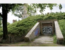Backsteingebäude mit gewölbtem Eingang, bedeckt von Graffiti, eingebettet in eine hügelige, von Bäumen und Sträuchern bewachsene Landschaft. Im Hintergrund ist ein hoher Backsteinturm erkennbar, teilweise verdeckt von Bäumen.