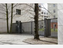 Hinterhof eines modernen Gebäudekomplexes mit einem hohen Metallzaun und einer breiten, geschlossenen Toranlage. Im Vordergrund stehen Bäume, und im Hintergrund sind Teile von Gebäuden mit cremefarbener und roter Fassade sichtbar.