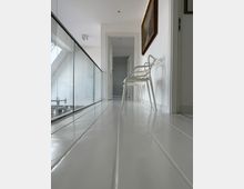 Moderner Flur mit hellen, weißen Wänden und glänzendem, weißem Boden. Links befindet sich ein Glasgeländer, rechts ein einzelner weißer Stuhl und ein gerahmtes Gemälde an der Wand; im Hintergrund ist eine geöffnete Tür zu sehen.