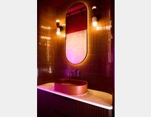 Ein modernes Badezimmer mit dunklen Fliesen an der Wand, einem ovalen, roten Waschbecken auf einer weißen, gesprenkelten Ablage und einem ovalen Spiegel mit integrierter Beleuchtung. Zwei Wandlampen befinden sich beidseitig des Spiegels, und die Szene ist in warmem, orange-pinkem Licht beleuchtet.