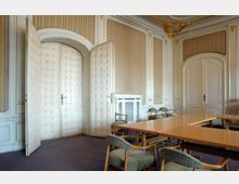 Ein klassisch gestalteter Innenraum mit gepolsterten weißen Doppeltüren, umgeben von verzierten Wänden mit goldenen Zierelementen. Im Vordergrund stehen Holztische und gepolsterte Stühle in einem Konferenz- oder Besprechungsraum.