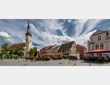 Ansicht eines historischen Marktplatzes mit einer Kirche im Hintergrund, Fachwerkhäusern und bunten Gebäuden auf der rechten Seite. Im Vordergrund befinden sich Café-Tische mit roten Sonnenschirmen und unterschiedlichem Stadtmobiliar.