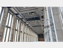 Innenansicht eines langen, teilsanierten Gebäudes mit hohen, rechteckigen Fenstern auf der linken Seite und einer abgehängten Decke mit geometrischen Deckenelementen. Die Wände sind teilweise freigelegte Ziegelmauerwerke, und freiliegende Stahlstrukturen sind sichtbar.