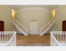 Ein heller Innenraum mit symmetrisch angeordneten, geschwungenen Treppen mit weißen Geländern, die auf beiden Seiten zu einer Doppeltür in der Mitte führen. Der Raum hat helle Wände, Holzböden und Türen aus dunklem Holz.