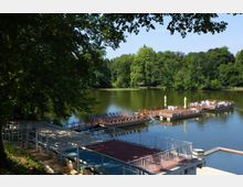 Lakeside recreation area with a calm green lake bordered by dense trees, and several large wooden floating docks with railings, seating, and sun umbrellas. In the foreground, stepped concrete terraces and metal railings lead down to the water.