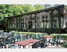 Außenansicht eines Gartenlokals mit Tischen und Stühlen auf einem Kiesplatz. Im Hintergrund eine historische, mit Efeu bewachsene Fassade mit großen Fenstern und einer überdachten Terrasse, umgeben von üppig grüner Natur.