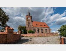 Backsteinkirche mit rotem Satteldach und einem Kirchturm, der eine Uhr trägt, umgeben von einer niedrigen Ziegelmauer. Im Vordergrund ist ein gepflasterter Weg und links sowie rechts kleinere Bäume und Laternen zu sehen, unter einer teils bewölkten Himmel.