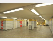 Unterirdischer U-Bahn-Bereich mit hell erleuchtetem, beige gefliestem Boden und Wänden in weiß-grauer Optik mit roten und blauen Elementen. Im Hintergrund führen Rolltreppen zu einer tiefergelegenen Ebene.