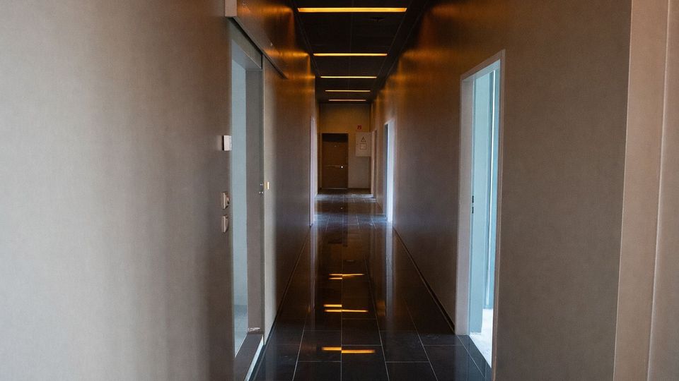 Langer, schmaler Innenflur in einem modernen Gebäude mit glatten grauen Wänden, mehreren Türöffnungen auf beiden Seiten und dunklem, glänzend gefliestem Boden. An der Decke verlaufen lineare Einbauleuchten, die sich im Boden spiegeln, am Ende des Flurs ist eine geschlossene Tür zu sehen.