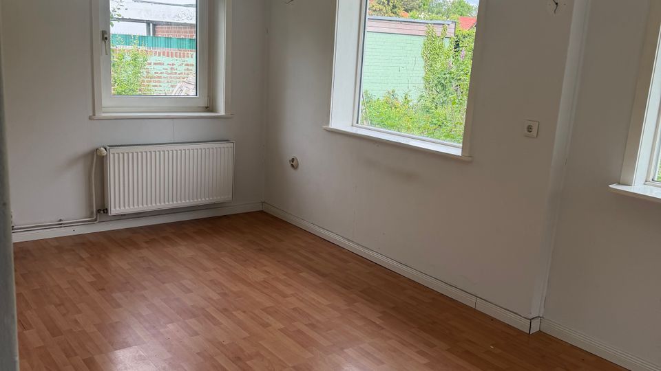 Leerer Wohnraum mit hellem Laminatboden, weißen Wänden und einem sichtbaren Holzbalken an der Decke. Zwei Fenster mit Blick ins Grüne sowie ein Heizkörper unter einem Fenster prägen die Raumseite.