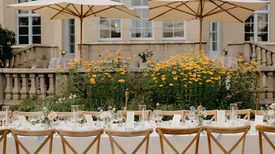 Lange, gedeckte Tafel mit weißen Tischdecken und Holzstühlen auf einer Rasenfläche, davor Blumenbeete und zwei große Sonnenschirme. Im Hintergrund steht ein helles, mehrstöckiges Herrenhaus mit großen Fenstern und einer steinernen Terrasse mit Balustrade.