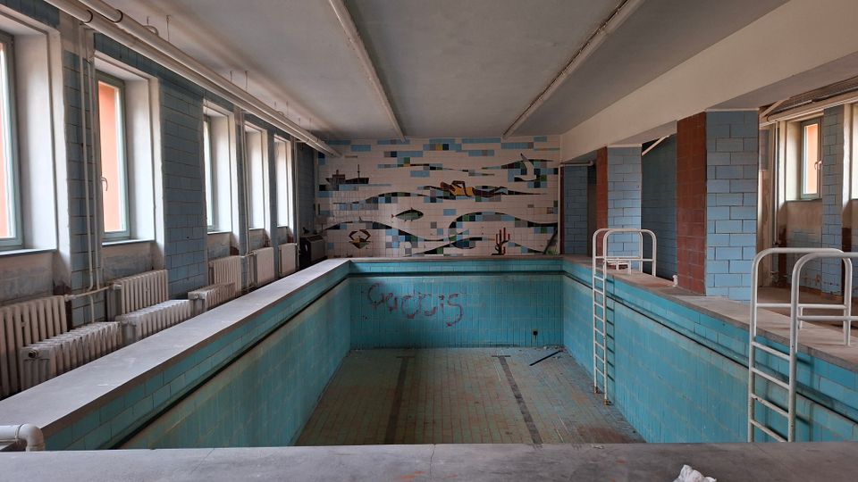 Leeres, gefliestes Hallenbad mit rechteckigem Becken, Metallleitern und umlaufendem Beckenrand. Links stehen hohe Fenster über Heizkörpern, an der Stirnwand ist ein Mosaikbild, und sichtbare Rohrleitungen verlaufen unter der Decke.