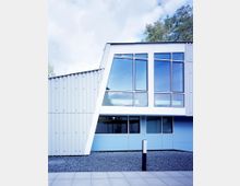 Modernes Gebäude mit wellenförmiger Metallfassade und großen Fenstern, die in einer weißen und blauen Konstruktion eingelassen sind. Der Vordergrund zeigt eine Fläche aus grauen Kieselsteinen und Pflasterplatten.