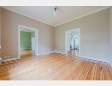 Leerer, heller Wohnraum mit breiten Holzdielen, hohen Decken mit Stuck und schlichtem Deckenlampenanschluss. Zwei große Türöffnungen mit weißen Rahmen führen in angrenzende Räume, einer mit grünen Wänden und einer mit Blick auf Fenster und Balkon.