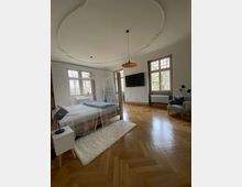 Ein geräumiges Schlafzimmer mit Fischgrätenparkett, großen Fenstern mit Sprossen, einem Doppelbett mit Baldachin, einem kleinen Nachttisch mit Lampe, einem Fernseher an der Wand und einem Sessel in einer Ecke. Die Decke hat ein dekoratives Stuckdetail, und der Raum ist hell durch das natürliche Licht.