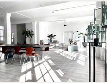 Heller, moderner Innenraum mit minimalistischer Einrichtung; ein langer, dunkler Tisch mit Stühlen in der Mitte, Lounge-Bereich mit Sofas und Pflanzen im Hintergrund sowie großen Fenstern, die Tageslicht einlassen.