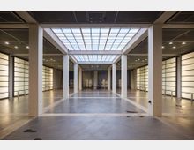 Ein moderner Innenraum mit hohen Decken und rechteckigen Säulen, die den Raum gliedern. Große Deckenfenster sowie seitliche, milchglasartige, beleuchtete Wände sorgen für gleichmäßige Ausleuchtung, während der Boden aus glänzendem Beton besteht.