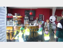 Ein kleines Café mit roter Wand, gelber Theke und schwarzen Barhockern. Im Hintergrund sind Regale mit Getränken und Snacks, eine Tafel mit Getränkepreisen sowie ein großer Kinoprojektor zu sehen. Im Vordergrund stehen Kinder in Warnwesten.