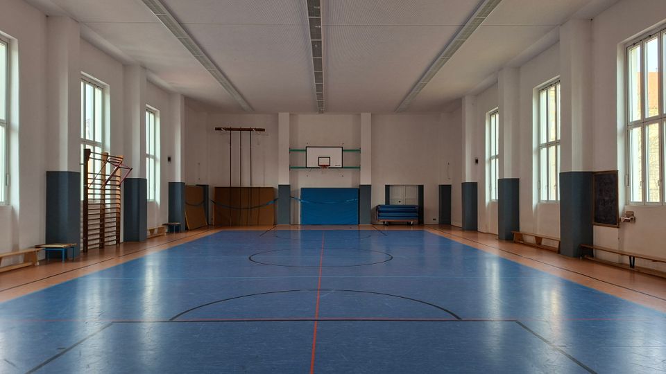 Leere Turnhalle mit blauem Sportboden und Markierungen, an der Stirnwand ein Basketballkorb mit Prallschutz. Hohe Fenster entlang der Seitenwände lassen Tageslicht herein; an den Wänden stehen Turngeräte, Matten und Bänke.