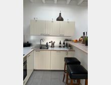 Kleine, moderne Küche mit cremefarbenen Schrankfronten, einer Edelstahlspüle und einer Arbeitsplatte aus Holz. Auf der Theke befinden sich zwei schwarze Barhocker, während verschiedene Küchenutensilien, Dekorationsgegenstände und Flaschen die Arbeitsflächen schmücken.