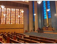 Innenraum einer modernen Kirche mit rechteckigen orangefarbenen und blauen Glasfenstern, die den Raum mit farbigem Licht durchfluten. Im Vordergrund befinden sich Holzbänke, während der erhöhte Altarbereich mit blauen Mosaikfliesen gestaltet ist und von schlichten Kerzen und violetten Paramenten dekoriert wird.