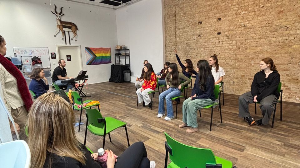 Interior of an open studio-style room with wood floors, a long exposed brick wall, and a grid ceiling with acoustic panels and hanging lights. Bright green chairs are arranged in rows facing a small setup at the front, with a pride flag and wall-mounted deer head decoration on the far wall.