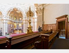 Innenraum einer Barockkirche mit reich verzierten Säulen, goldenen Dekorationen und Kirchenbänken aus dunklem Holz. Im Hintergrund sind ein kunstvoll gestalteter Altar, bunte Kirchenfenster und eine große Kuppeldecke zu sehen.