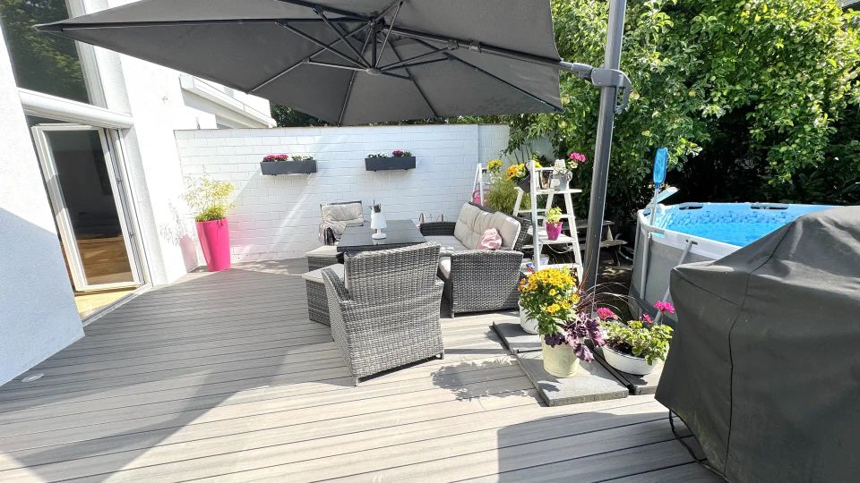Holzterrasse an einem Wohnhaus mit großem Ampelschirm, Rattan-Sitzgruppe und Pflanzkübeln vor einer weißen Mauer mit Blumenkästen. Rechts stehen ein aufstellbarer Pool neben dichtem Grün sowie ein abgedeckter Grill.