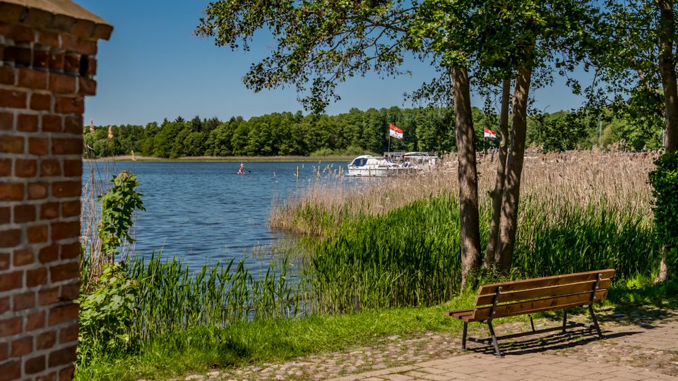 Uferweg an einem See mit Schilfgürtel, Bäumen und einer Holzbank im Vordergrund; links ist ein Ziegelmauerstück zu sehen. Auf dem Wasser liegen mehrere Boote nahe einem Steg, im Hintergrund verläuft eine bewaldete Uferlinie unter blauem Himmel.