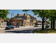 Bahnhofsgebäude aus gelbem Backstein mit arkadenartigen Fenstern, flankiert von Bushaltestellen und einem Parkplatz im Vordergrund. Bäume spenden Schatten, ein geparkter PKW und ein Linienbus sind sichtbar.