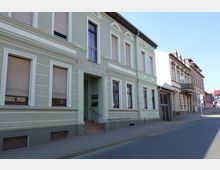 Straßenszene mit einer Reihe von Wohnhäusern im Altbaustil, teils mit verputzten Fassaden und dekorativen Fensterrahmen. Der Gehweg aus Pflastersteinen grenzt an eine asphaltierte Straße, und das Bild zeigt eine ruhige Stadtumgebung bei klarem Himmel.