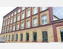 Fassade eines mehrstöckigen Gebäudes aus gelblichen Ziegelsteinen mit roten Backsteinelementen. Die Wände sind regelmäßig durch hohe, weiße Fenster mit Rundbögen unterteilt.