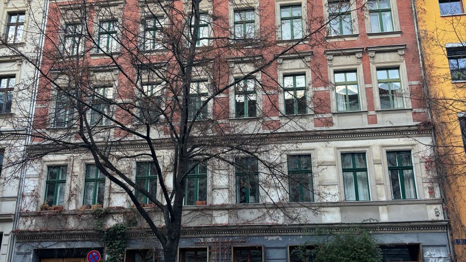 Mehrstöckiges Altbau-Wohnhaus mit rot-weiß gegliederter Fassade und vielen rechteckigen Fenstern, gesehen von der Straße aus. Im Vordergrund stehen ein kahler Baum sowie parkende Autos und Fahrräder; im Erdgeschoss sind graffitibedeckte Eingänge und Schaufenster zu sehen.