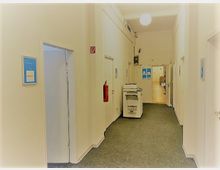 Ein heller Büroflur mit weißen Wänden und grauem Teppichboden. Links und rechts befinden sich Türen, in der Mitte steht ein Kopierer, und am Ende des Flurs sind weitere Räume sichtbar.