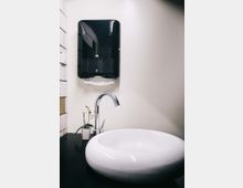 Weißes Badezimmerwaschbecken mit verchromtem Wasserhahn auf einer schwarzen Arbeitsfläche, darüber ein schwarzer Papierhandtuchspender an einer cremefarbenen Wand; daneben steht eine kleine Orchidee in einem weißen Topf.