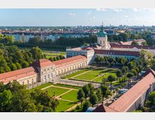 Luftaufnahme eines Barockschlosses mit einer markanten Kuppel, flankiert von langgestreckten Seitenflügeln mit roten Dächern und großen Fensterfronten. Im Vordergrund sind symmetrische Gartenanlagen mit Rasenflächen, Gehwegen und Bäumen zu sehen, während die Umgebung von einer Stadtlandschaft mit Wohnhäusern umgeben ist.