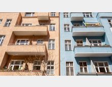 Zwei benachbarte Wohngebäude mit Putzfassaden, die linke in einem beige-orangenen Ton und die rechte in Blau. Beide Gebäude haben mehrere Balkone und weiße Fensterrahmen, ergänzt durch Pflanzen und gelüftete Fenster.