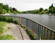 Flussufer mit einer geschwungenen Metallgeländerabsperrung, angrenzend an Grasflächen und Büsche. Im Hintergrund sind Bäume, Boote an einem Steg und vereinzelte Häuser zu sehen.