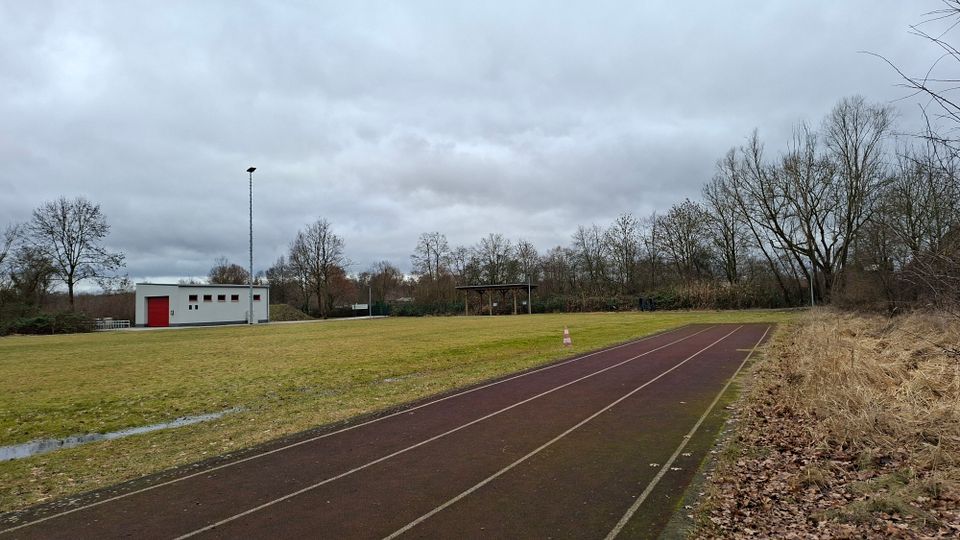 Leichtathletikanlage im Freien mit Laufbahn entlang einer großen Rasenfläche, daneben ein kleiner Funktionsbau mit rotem Tor und ein hoher Flutlichtmast. Im Hintergrund stehen ein überdachter Unterstand und eine Baumreihe unter bedecktem Himmel.