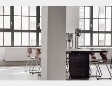 Beleuchteter Besprechungsraum mit modernen Stühlen und einem langen Tisch aus Holz, umgeben von großen Fenstern mit schwarzen Rahmen, durch die Tageslicht einfällt. Im Hintergrund sind Heizkörper unterhalb der Fenster sichtbar, ergänzt durch schlichte Bodenleuchten.