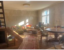 Wohnzimmer mit einer Mischung aus Retro- und Vintage-Möbeln, darunter eine grüne Polstercouch, ein runder Holztisch, mehrere Stühle und kleinere Tische. Der Raum hat eine helle, lichtdurchflutete Atmosphäre, unbehandelte Wände, Holzboden, einen orientalischen Teppich und ein großes Fenster, durch das Sonnenlicht einfällt.