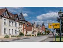 Straßenszene in einer Kleinstadt mit historischen Wohnhäusern im Stil des 19. Jahrhunderts, gekennzeichnet durch Verzierungen und spitze Dächer. Rechts ist ein gelbes Verkehrsschild mit Richtungsangaben zum "Zentrum" und nach "Wittenberge" zu sehen, umgeben von grünen Bäumen und Straßenlaternen.