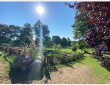 Sonniger Park mit grünem Rasen, vereinzelten Bäumen und einem kleinen, abgezäunten Gartenbereich aus Holzpfählen. Im Vordergrund liegen große Steine, umgeben von Blumen, während der helle Himmel und ein strahlender Sonnenstrahl die Szene erhellen.